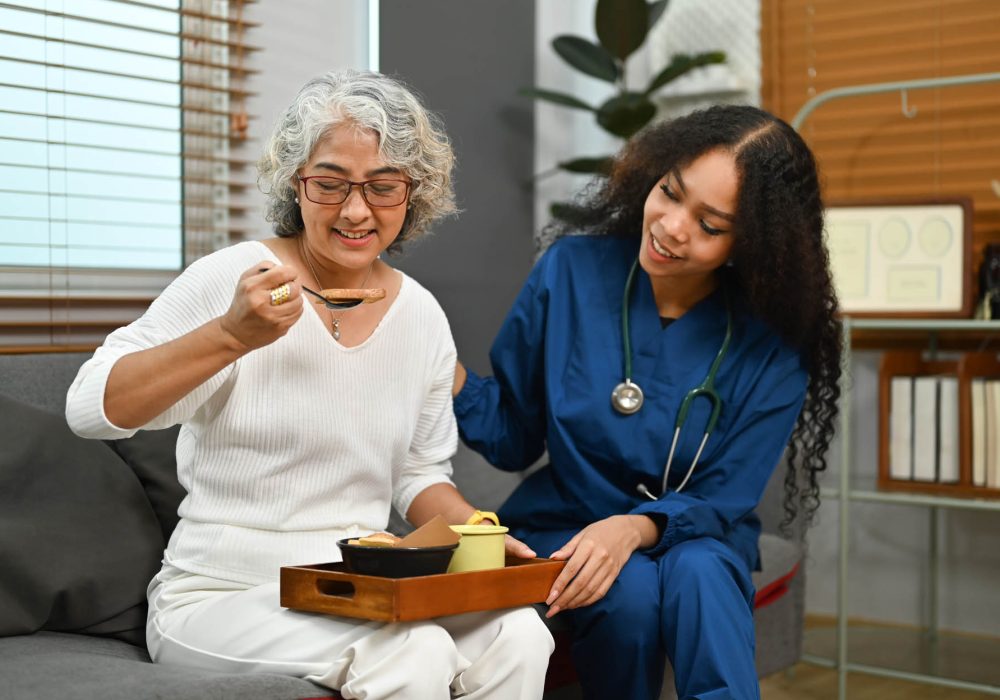 Photo of elderly woman having healthy nutrition breakfast meal with professional helpful caregiver on couch in the morning.